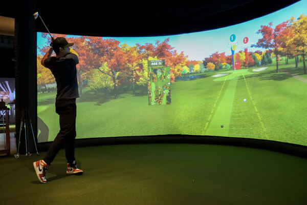 Golfer mid-swing in front of a Tee and Tap simulator impact screen showing a bright fairway
